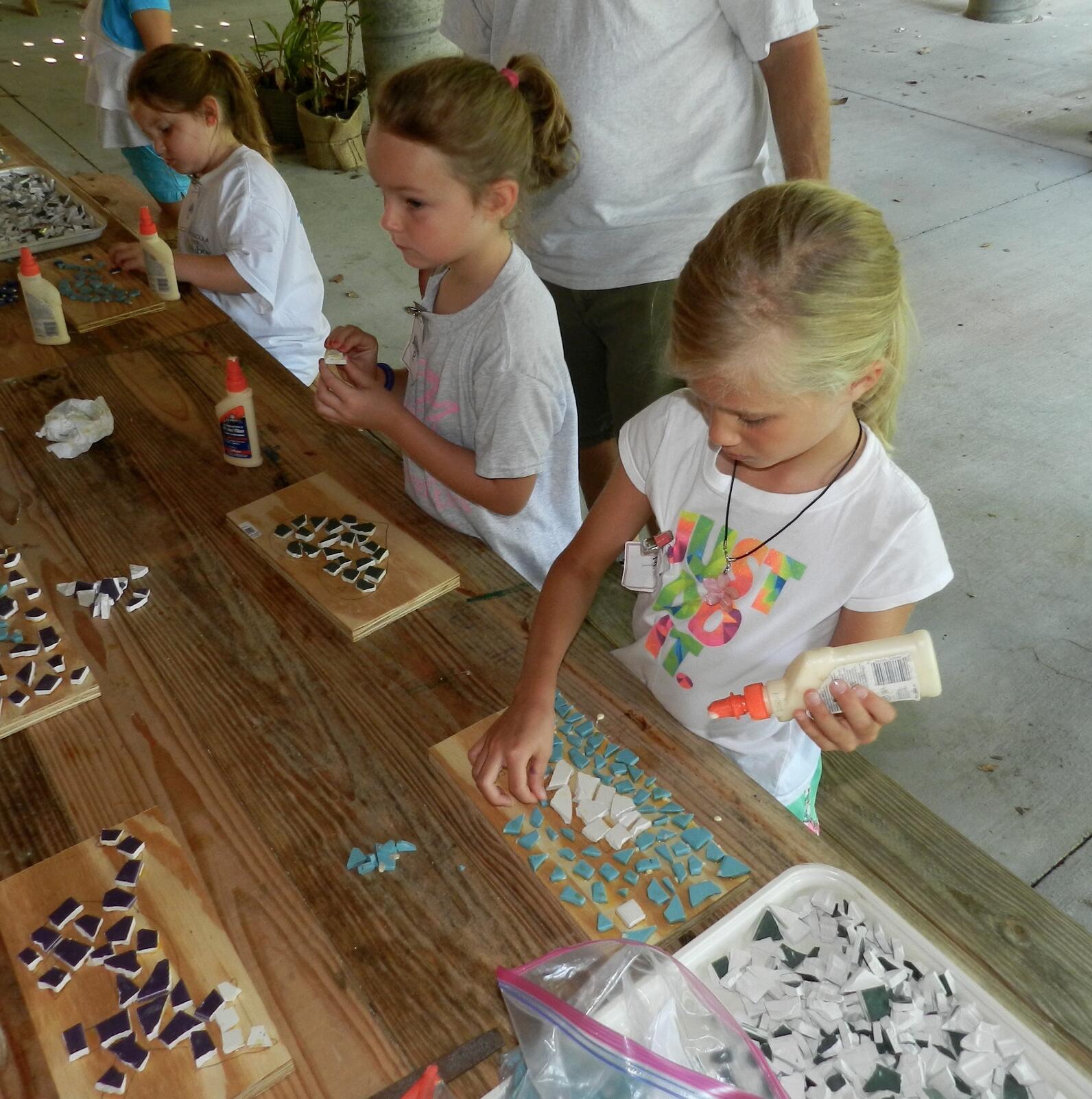 Three young girls use liquid glue and broken glass to create a mosaic on a slab of wood.