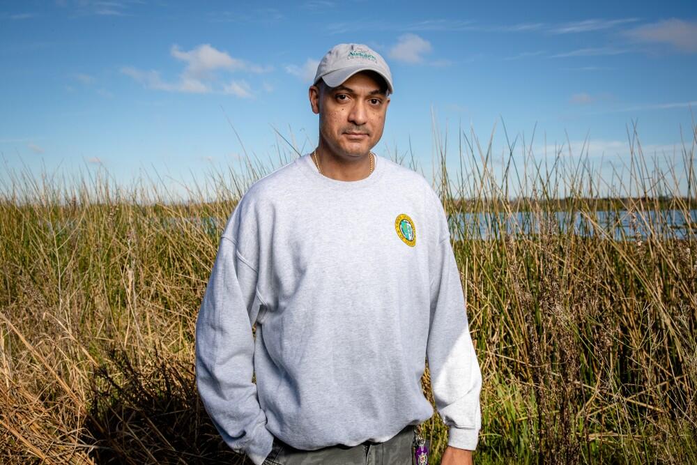 A man wearing an Audubon baseball cap and a grey crewneck sweater stands in front of a lake bordered by tall grass.