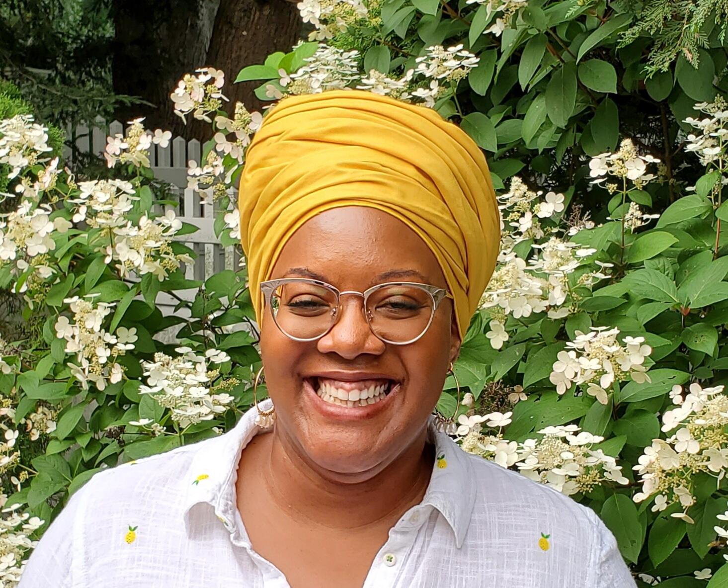 A woman wearing glasses, a white collared shirt with printed lemons, and a yellow head wrap.