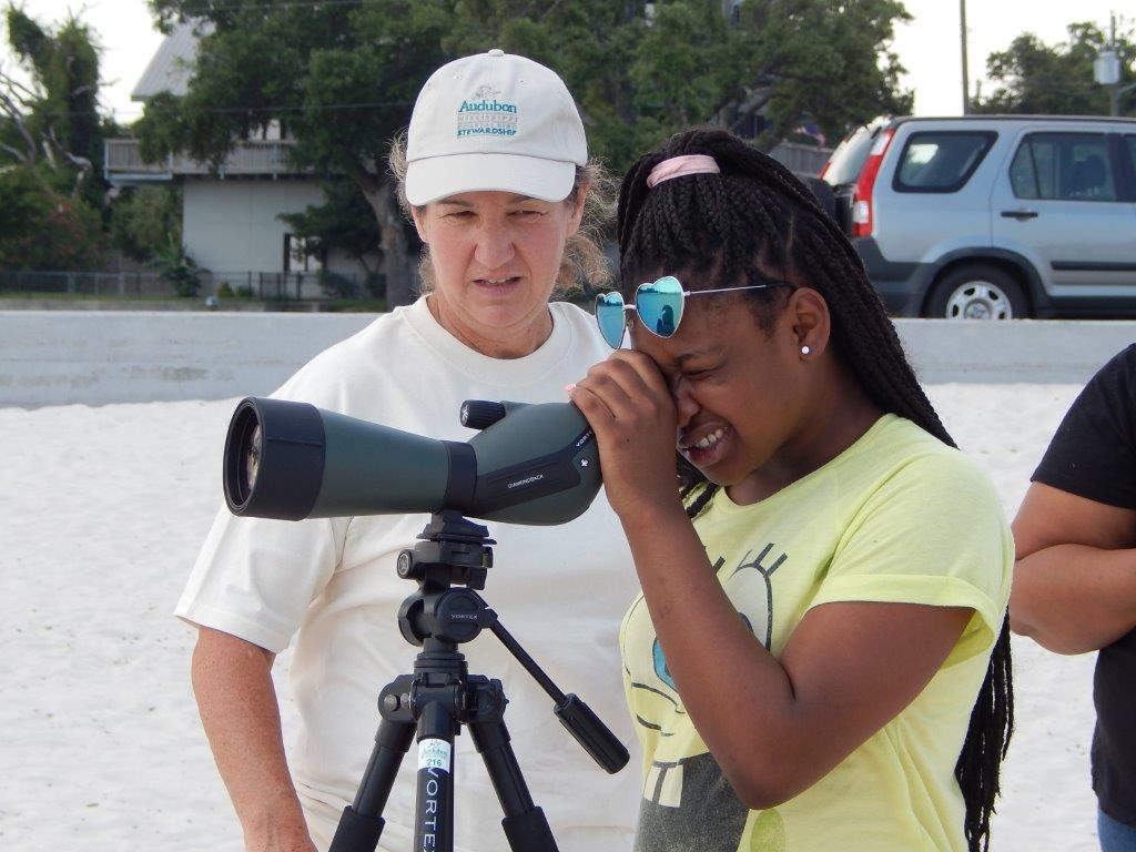 A young girl wearing a Spongebob Squarepants t-shirt and heart shaped sunglasses looks through a scope. A woman wearing an Audubon stewardship hat stands behind her.