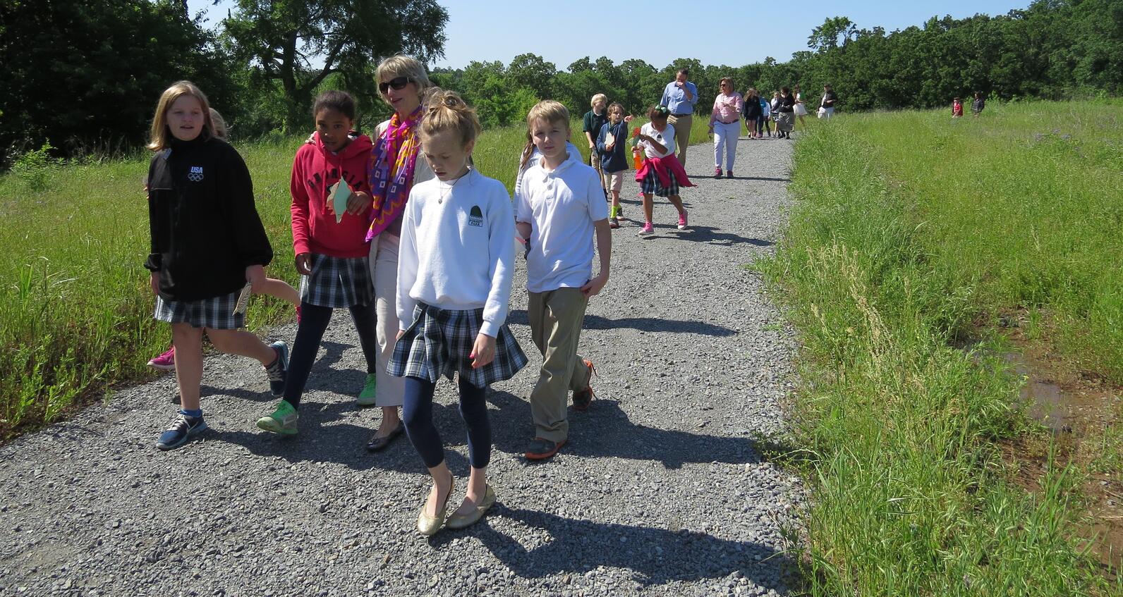 A group of school kids and teachers walk along a gravel path carved through tall grass.