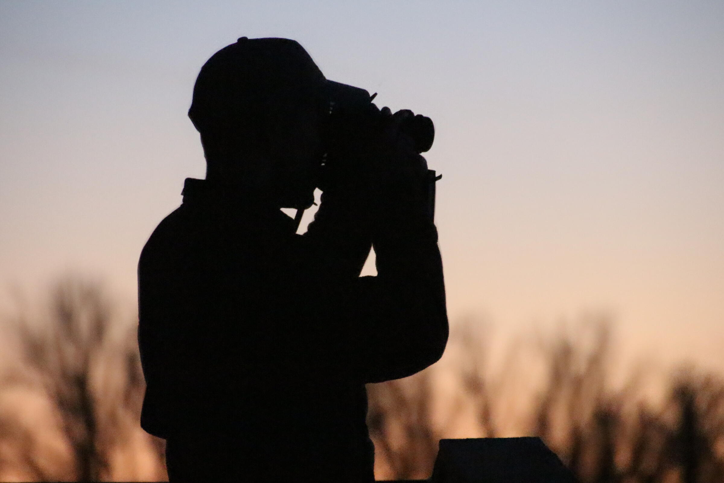 A birder wearing a baseball cap and looking through binoculars is backlit by a blue and pink sunset.