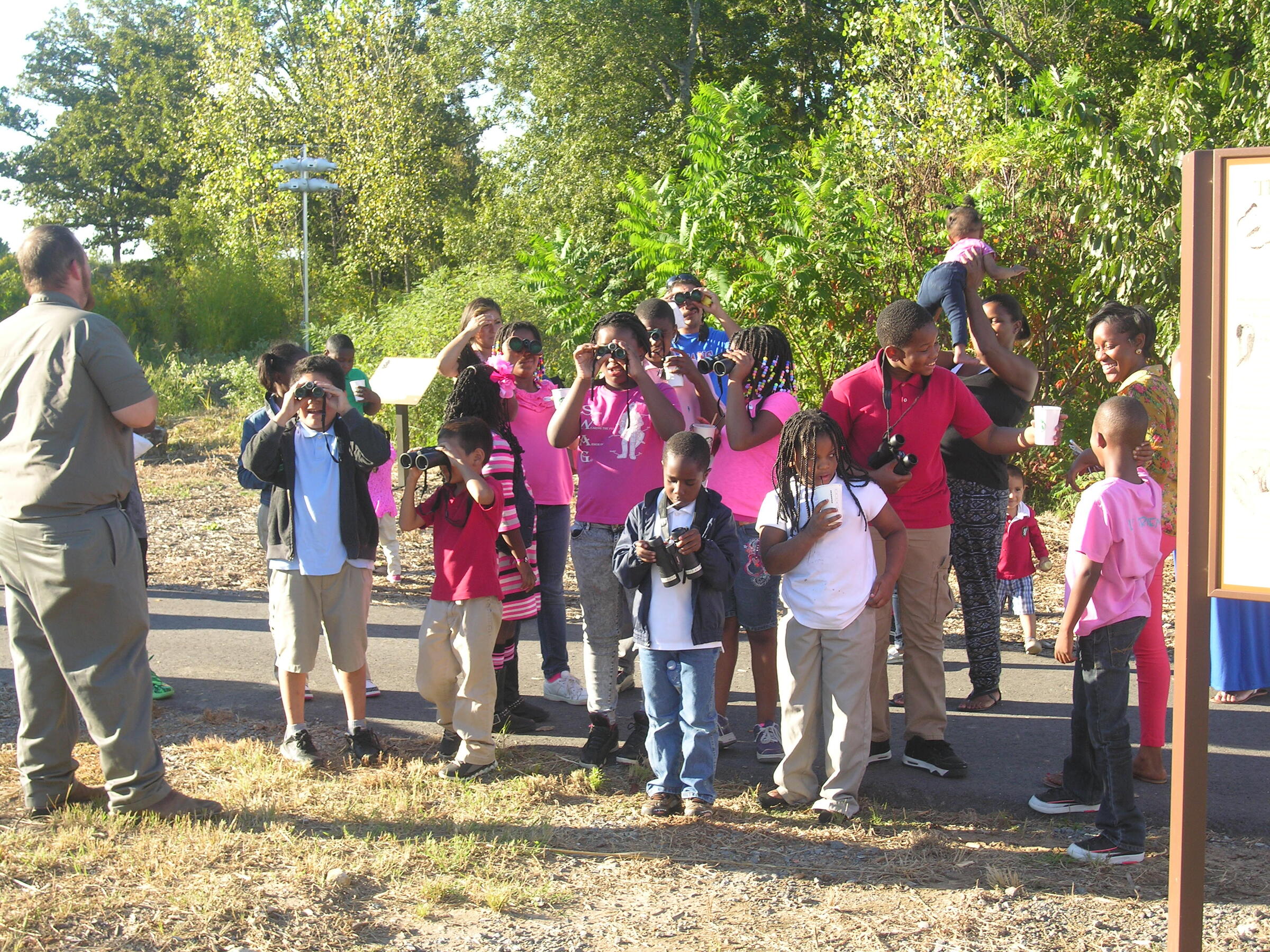 A group of kids and adults gather on a path while looking through binoculars.